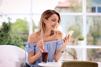 Photo of Young woman using mobile phone while drinking tasty healthy smoothie at table, indoors