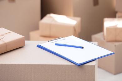 Photo of Clipboard, cardboard box and blurred stacked parcels on background, indoors