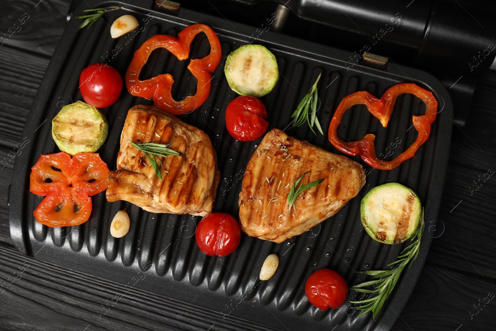 Photo of Electric grill with tasty meat, spices and vegetables on black wooden table, top view