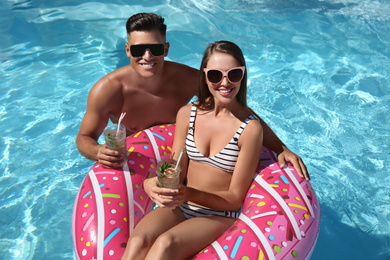 Photo of Happy couple with inflatable ring and cocktails in outdoor swimming pool on sunny summer day