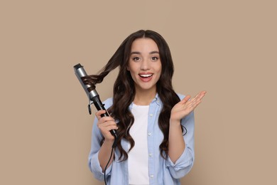 Photo of Excited woman using curling hair iron on beige background