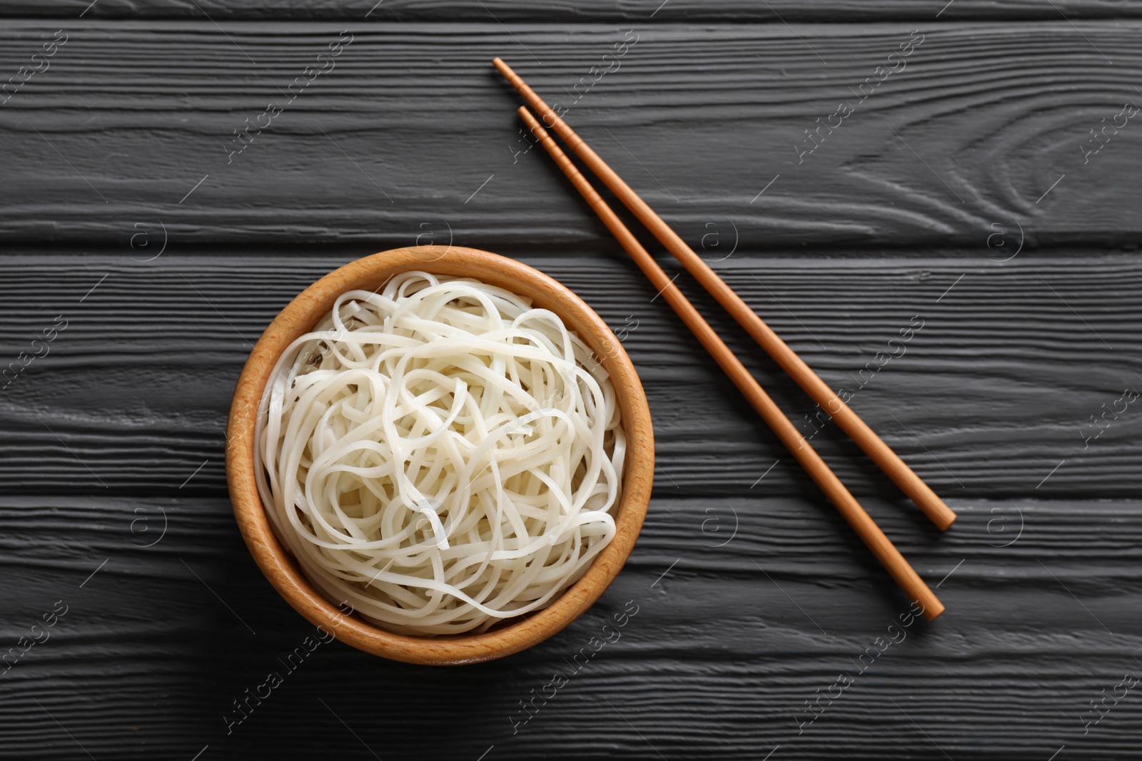 Photo of Bowl of tasty cooked rice noodles and chopsticks on black wooden table, flat lay