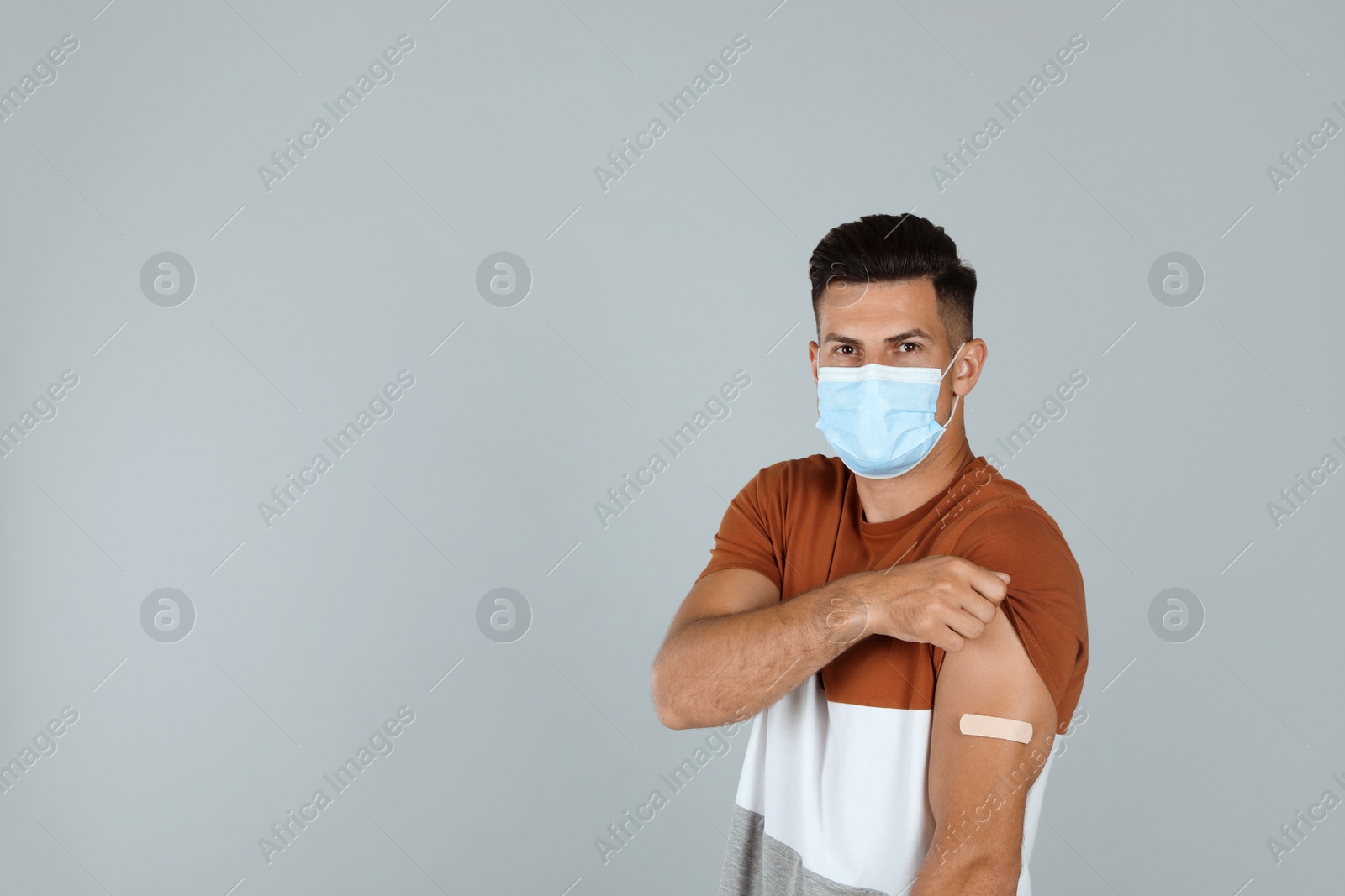 Photo of Vaccinated man with protective mask showing medical plaster on his arm against grey background. Space for text