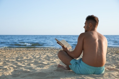 Young man reading book on sandy beach near sea