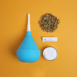 Enema, suppository, dry chamomile and bowl of salt on yellow background, flat lay