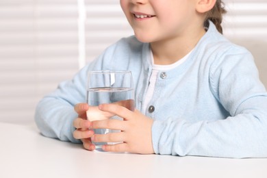 Photo of Little girl holding glass of fresh water at white table indoors, closeup