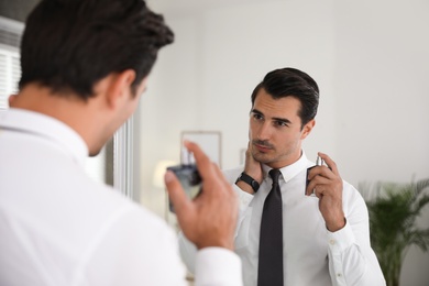 Handsome young man using perfume near mirror indoors