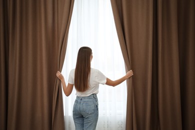 Woman opening elegant window curtains in room, back view