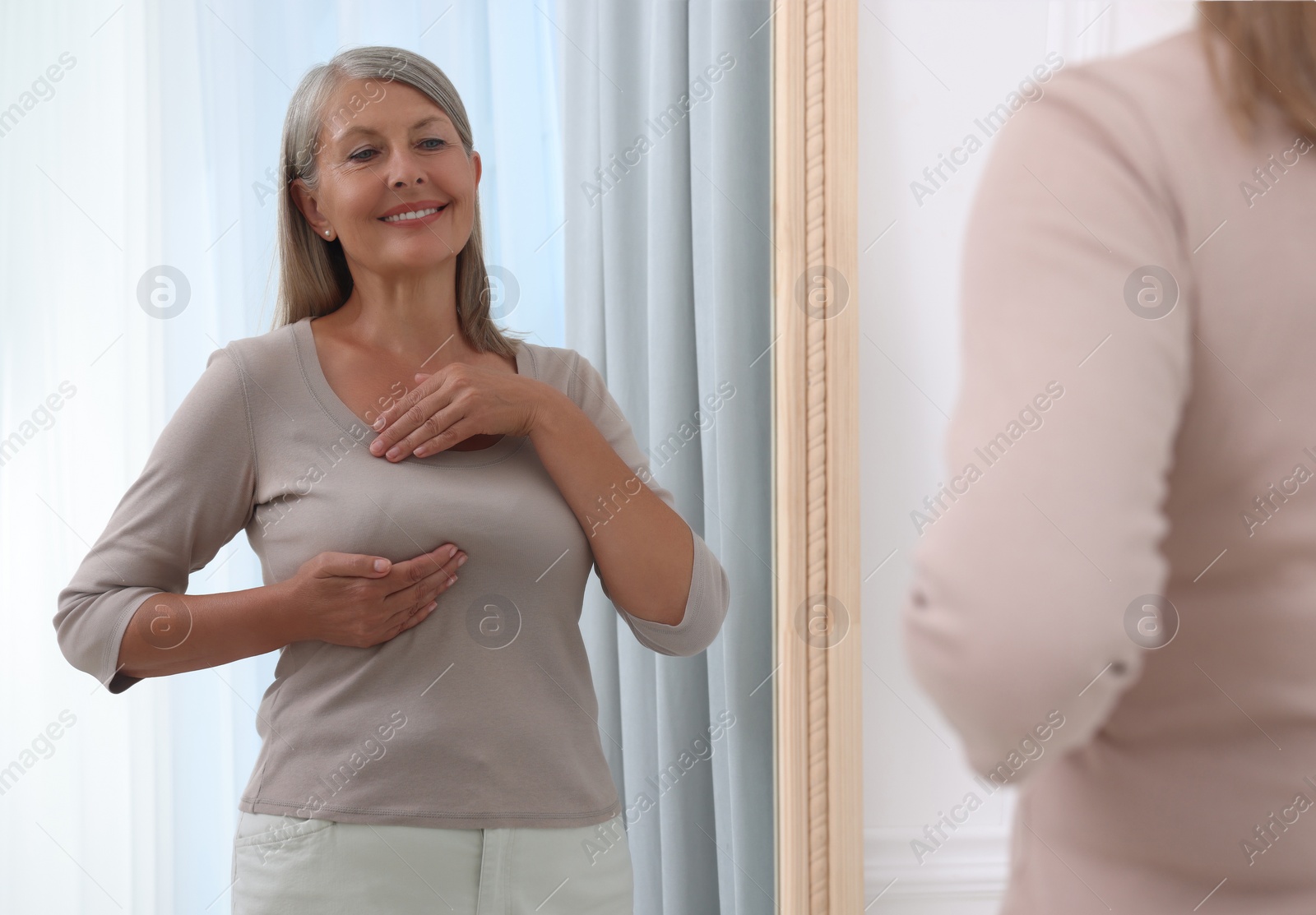 Photo of Beautiful senior woman doing breast self-examination near mirror indoors