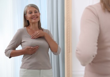 Beautiful senior woman doing breast self-examination near mirror indoors