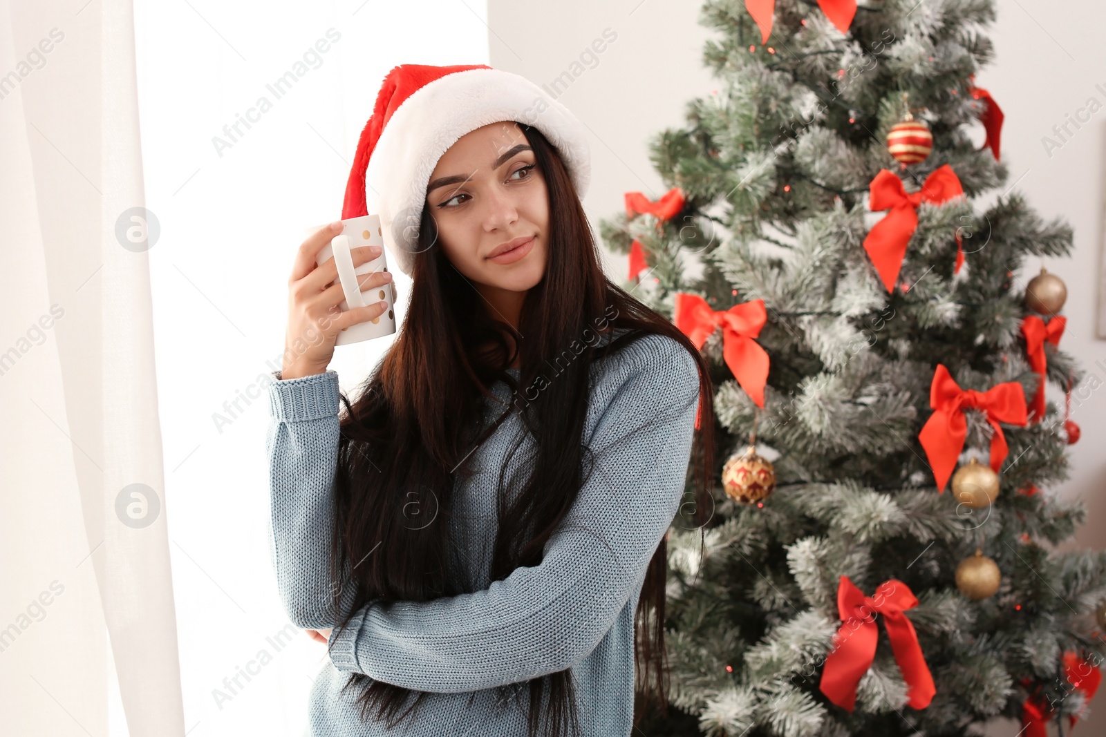 Photo of Beautiful young woman in Santa hat with cup of drink near Christmas tree at home