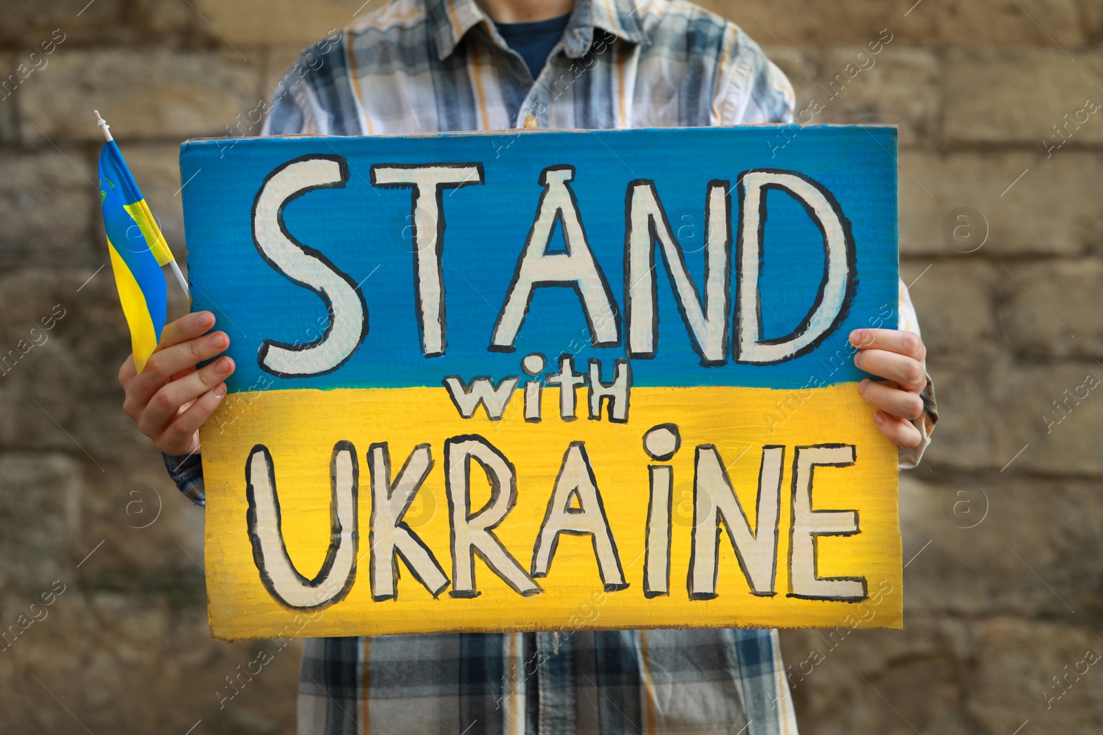 Photo of Woman holding poster in colors of national flag and words Stand With Ukraine against brick wall outdoors, closeup