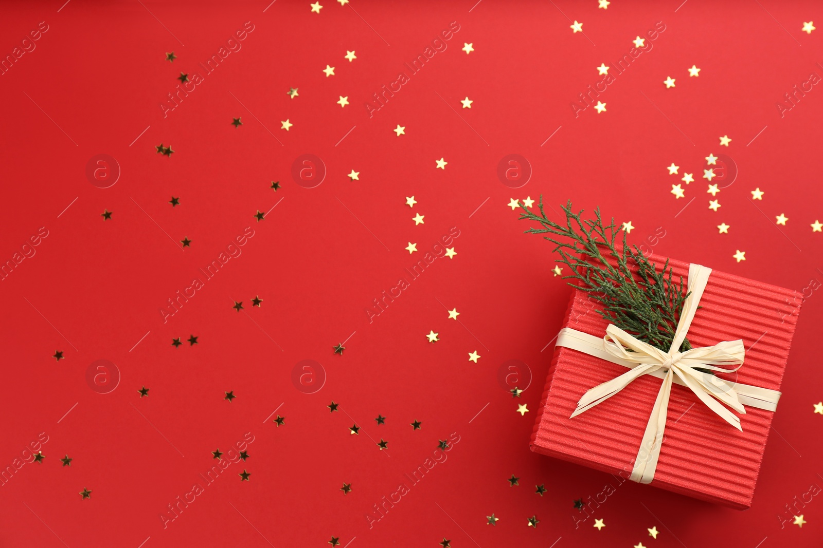 Photo of Christmas gift box with coniferous twigs and shiny confetti on red background, flat lay. Space for text