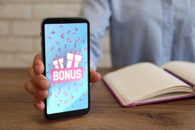 Image of Bonus gaining. Woman showing smartphone at wooden table, closeup. Illustration of gift boxes, word and falling confetti on device screen