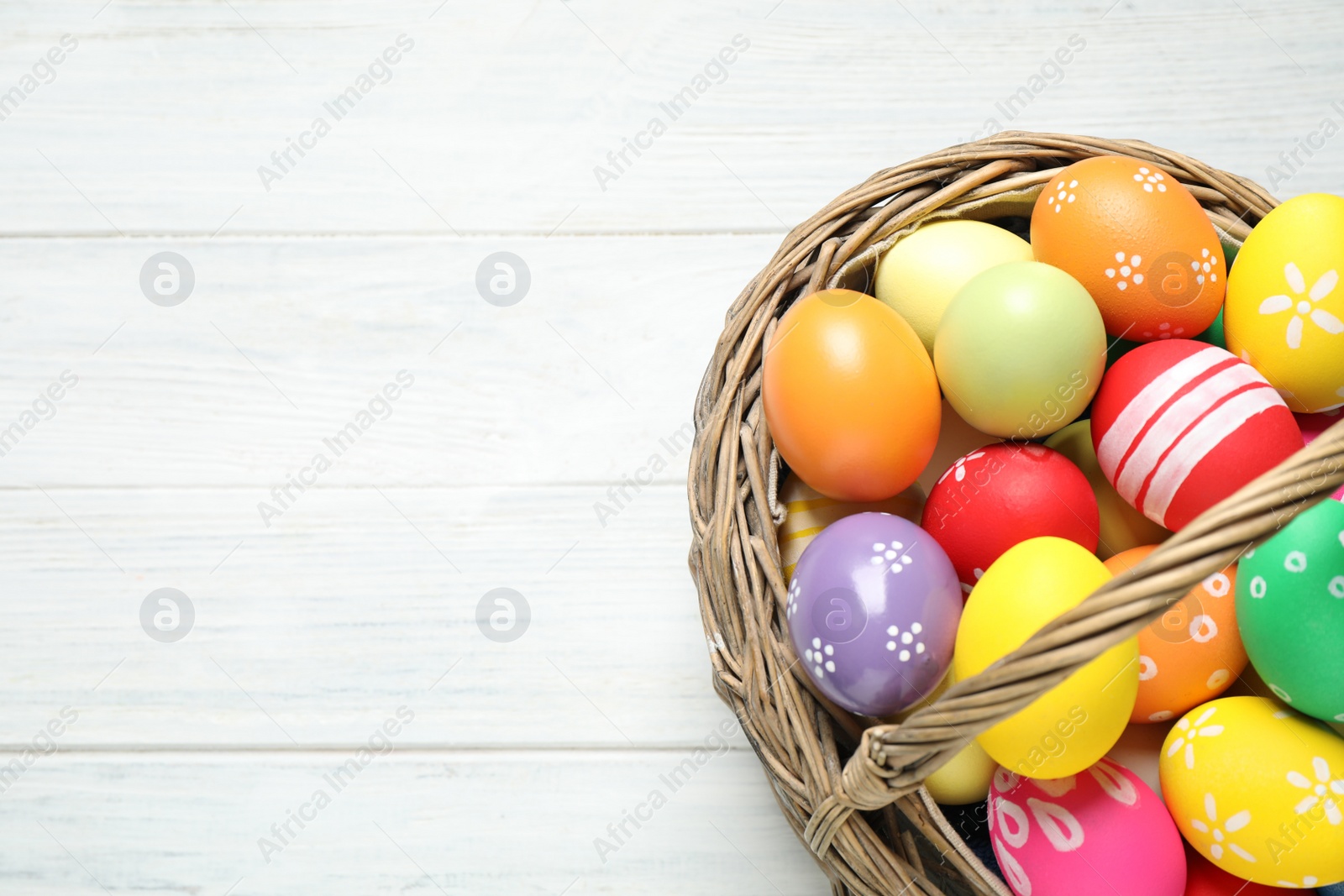 Photo of Colorful Easter eggs in basket on white wooden table, top view. Space for text