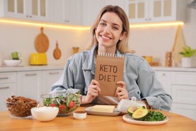 Photo of Woman holding notebook with phrase Keto Diet and different products at wooden table in kitchen