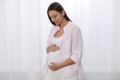 Beautiful pregnant woman in pink shirt near window indoors