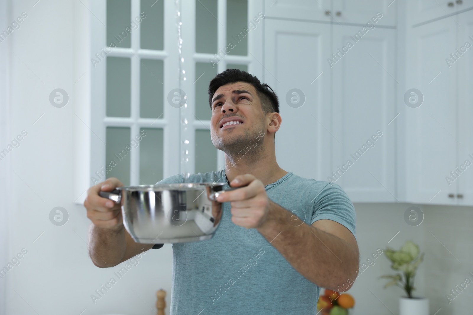 Photo of Emotional man collecting water leaking from ceiling in kitchen. Damaged roof