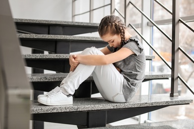 Photo of Depressed little girl sitting on stairs indoors. Time to visit child psychologist