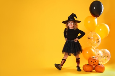 Photo of Cute little girl with pumpkins and balloons wearing Halloween costume on yellow background. Space for text