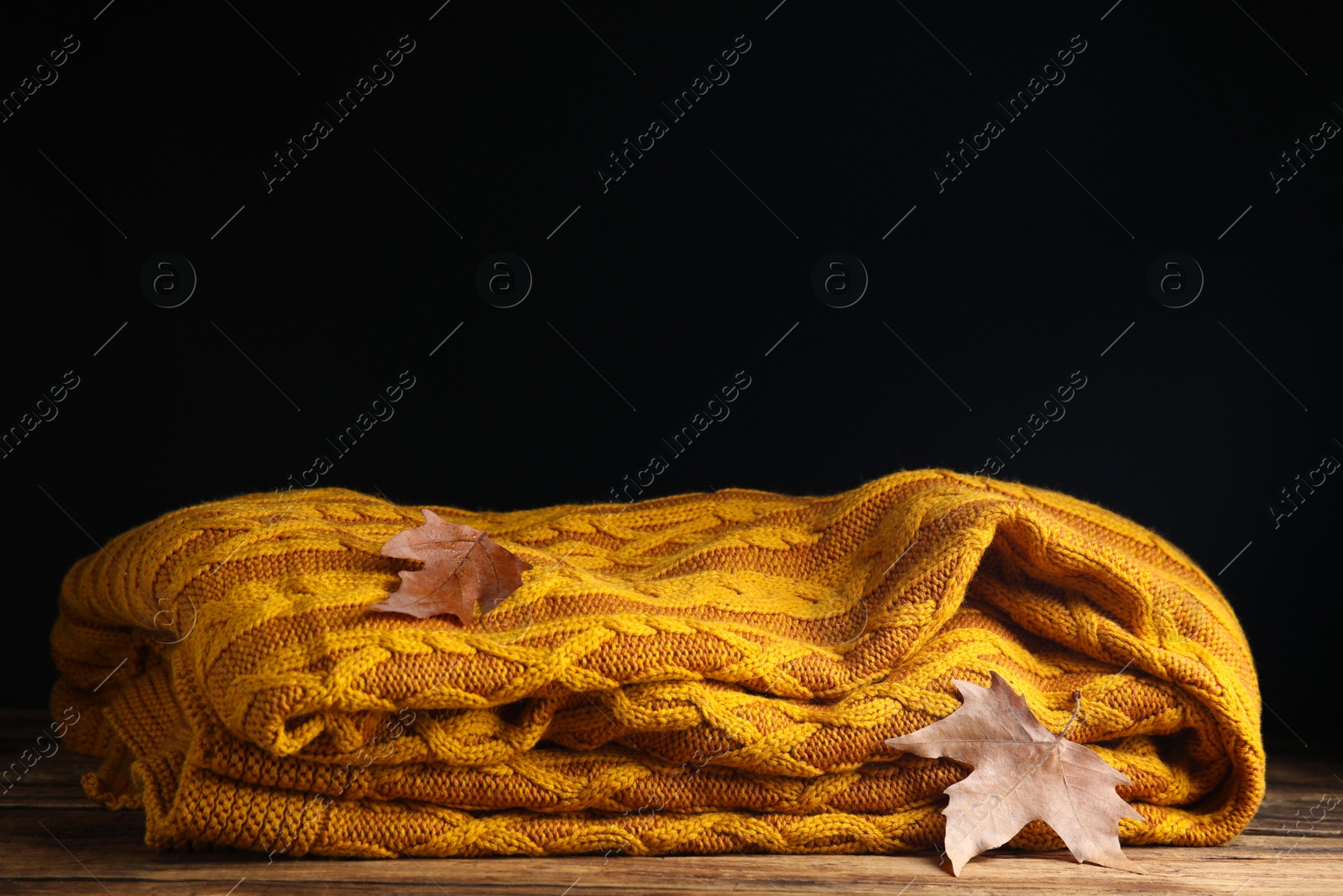 Photo of Orange knitted plaid with dry leaves on wooden table. Space for text