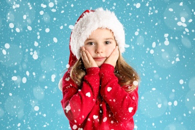 Image of Cute child in Santa hat under snowfall on light blue background. Christmas celebration