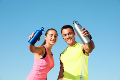 Young sporty couple with water bottles against blue sky on sunny day