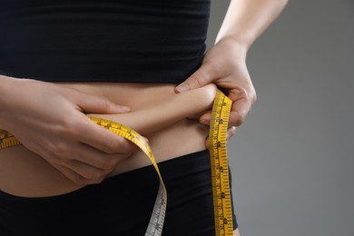 Photo of Woman with measuring tape touching belly fat on grey background, closeup. Overweight problem