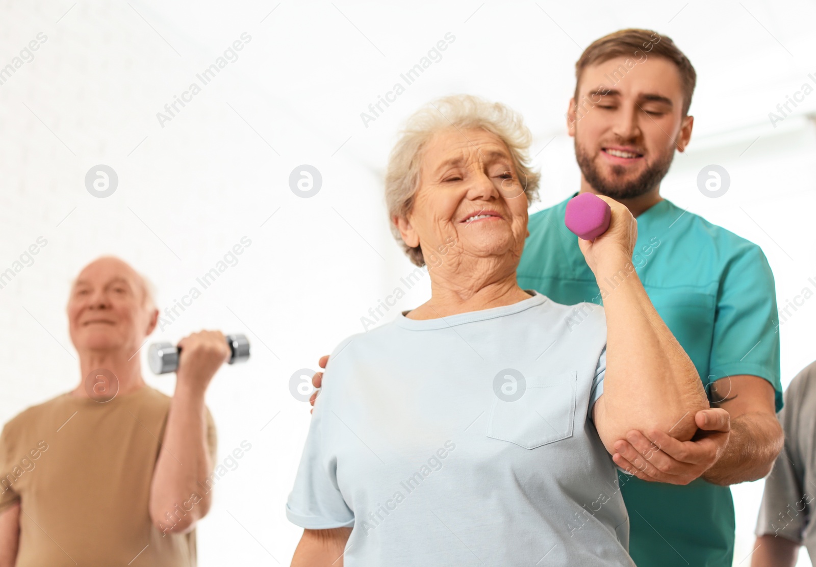 Photo of Care worker helping elderly woman to do exercise with dumbbell in hospital gym