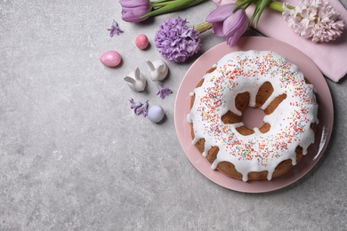Photo of Glazed Easter cake with sprinkles, painted eggs and flowers on grey table, flat lay. Space for text