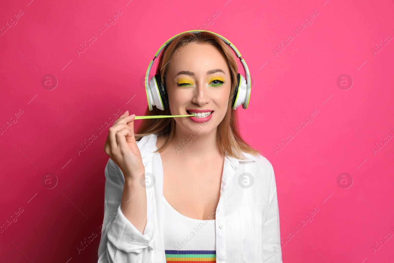 Photo of Fashionable young woman with bright makeup and headphones chewing bubblegum on pink background