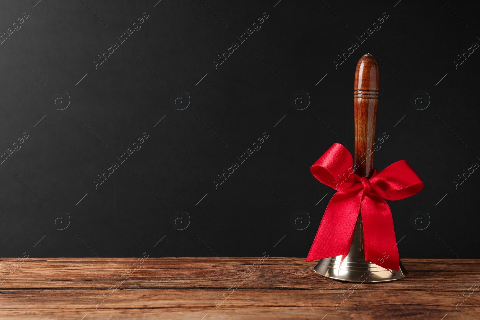 Photo of Golden bell with red bow on wooden table near blackboard, space for text. School days