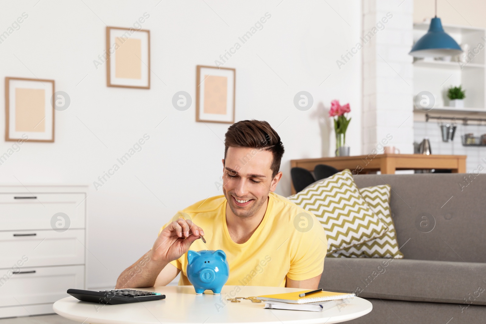 Photo of Happy man putting coin into piggy bank at table in living room. Saving money