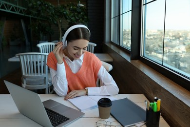 Photo of Young female student with laptop and headphones studying at table in cafe