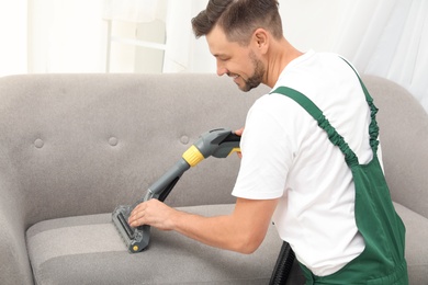 Male janitor removing dirt from sofa with upholstery cleaner indoors