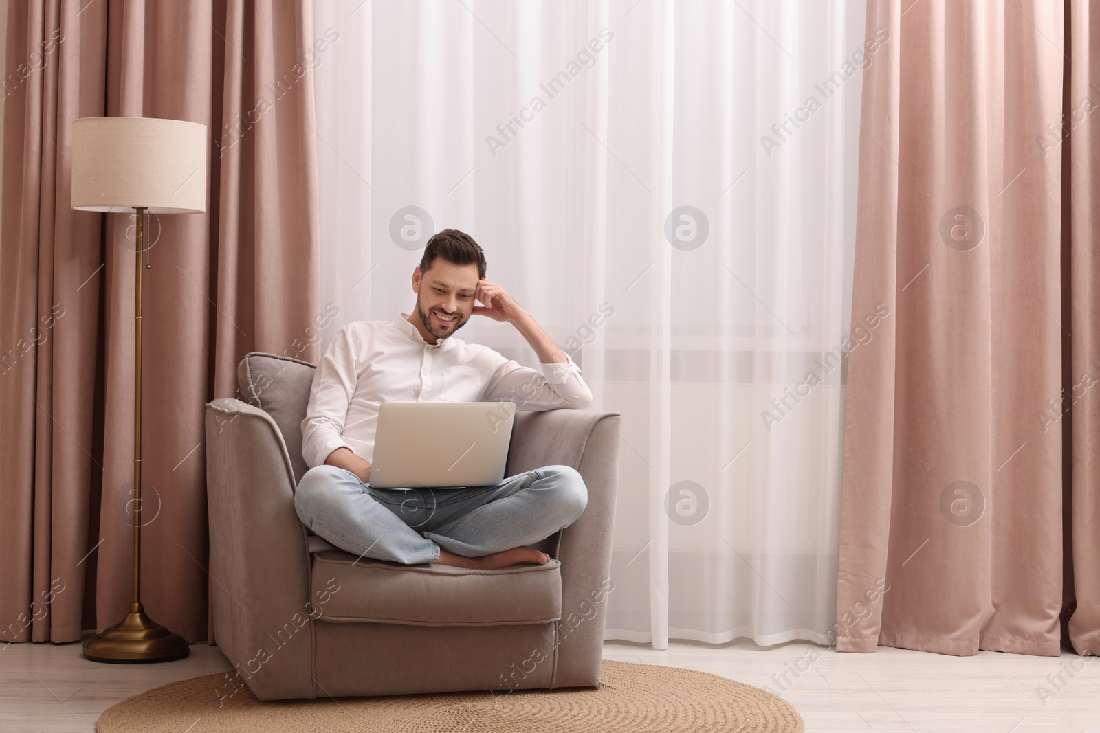 Photo of Smiling man holding laptop on armchair near window with beautiful curtains at home. Space for text