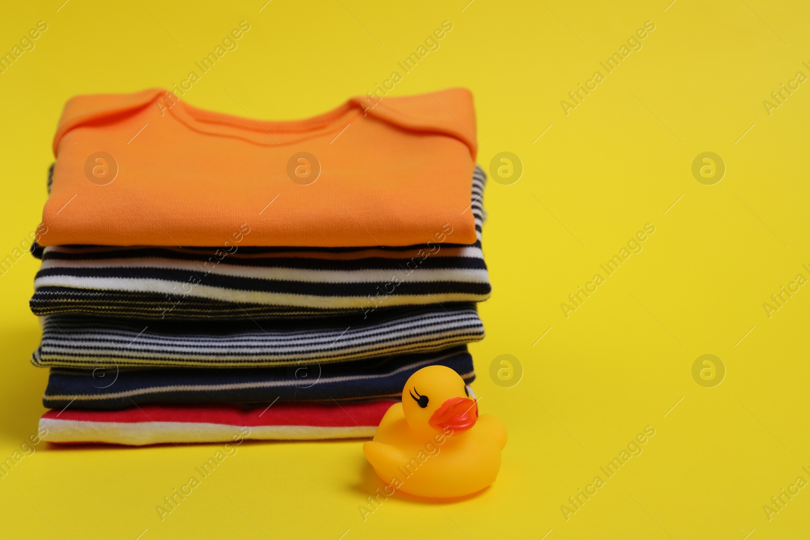 Photo of Stack of clean baby clothes and rubber duck on yellow background. Space for text