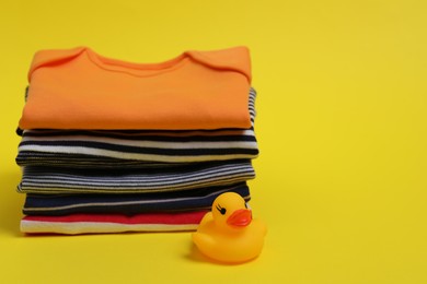 Photo of Stack of clean baby clothes and rubber duck on yellow background. Space for text