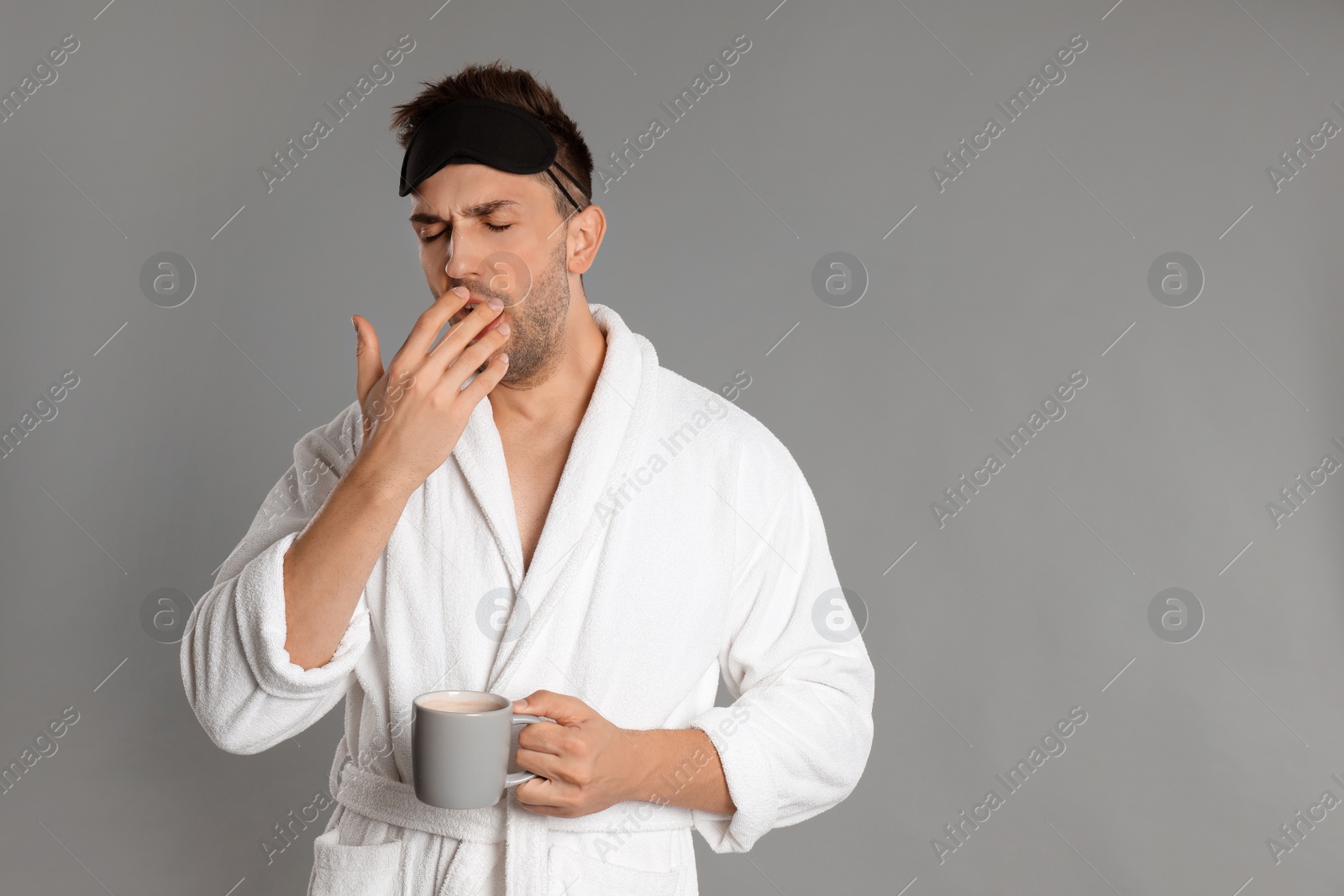 Photo of Young man in bathrobe yawning on grey background, space for text