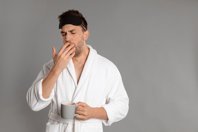 Young man in bathrobe yawning on grey background, space for text