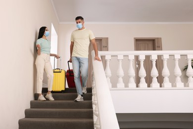 Photo of Couple wearing medical face masks with suitcases going down stairs in hotel