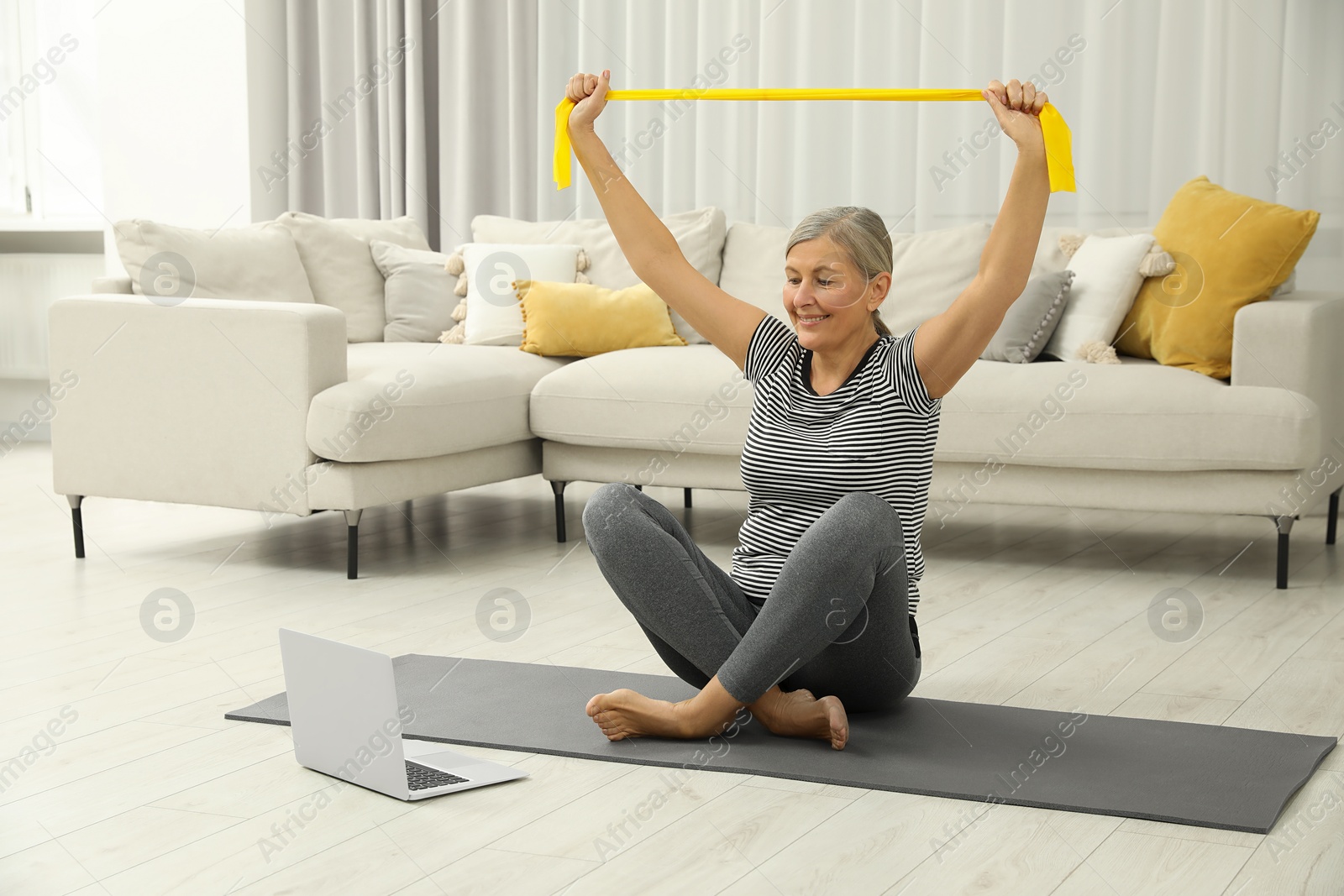 Photo of Senior woman doing exercise with fitness elastic band near laptop on mat at home