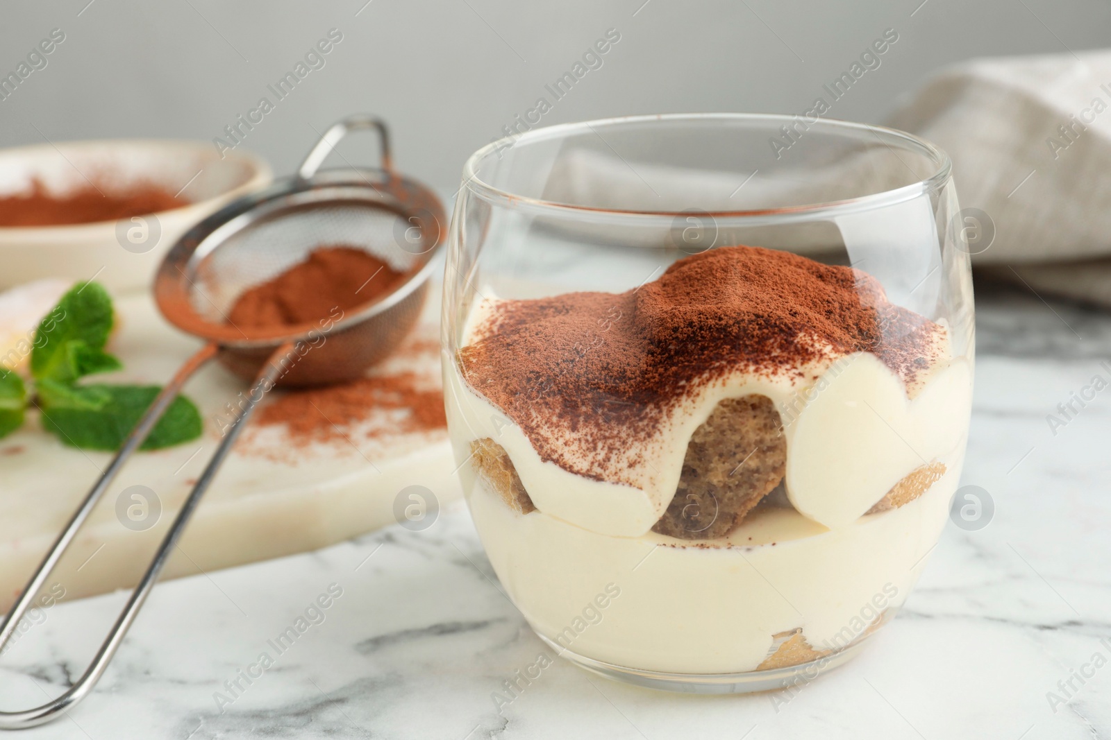 Photo of Delicious tiramisu in glasses and sieve with cocoa powder on white marble table, closeup