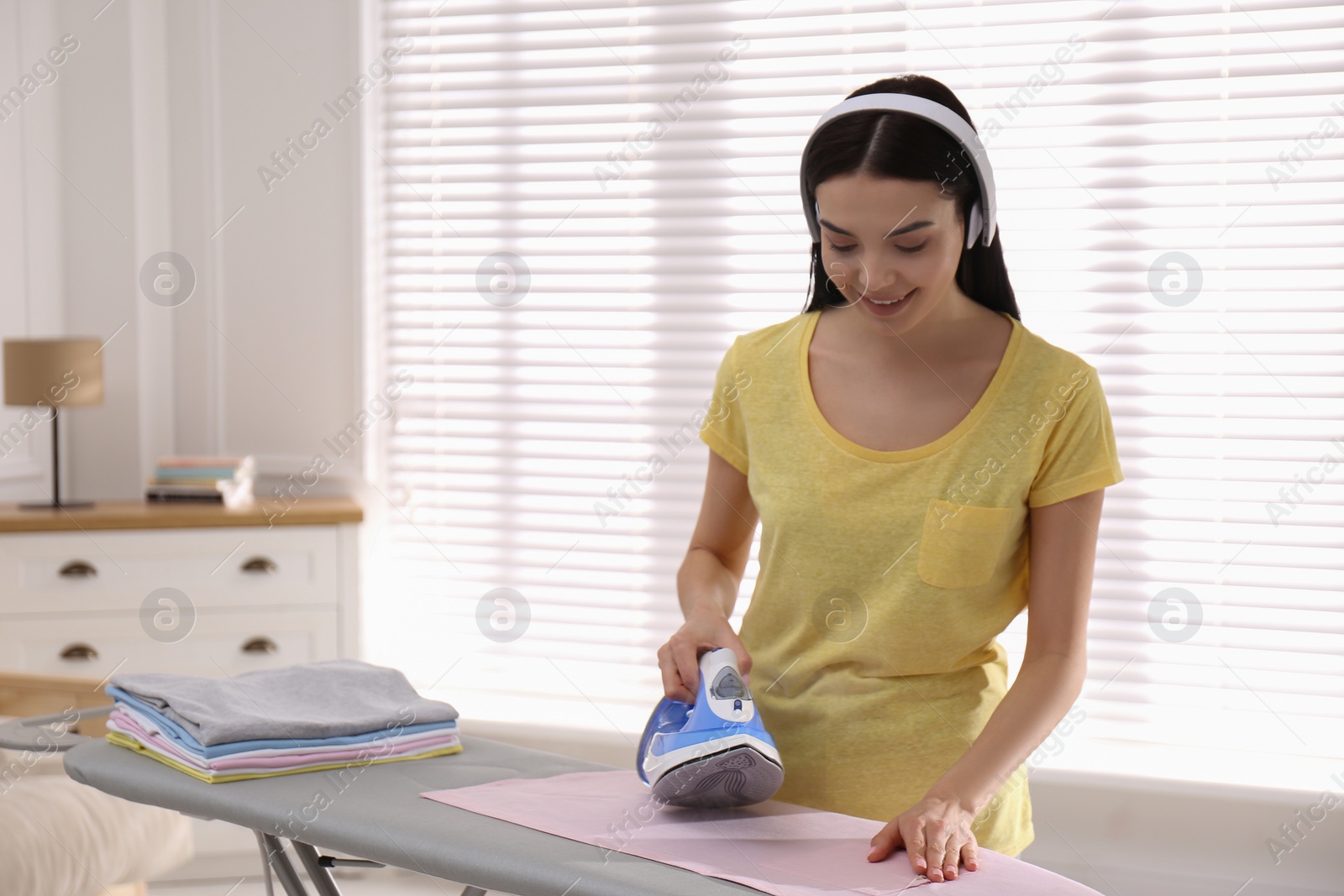 Photo of Young woman listening to music while ironing clothes at home. Space for text