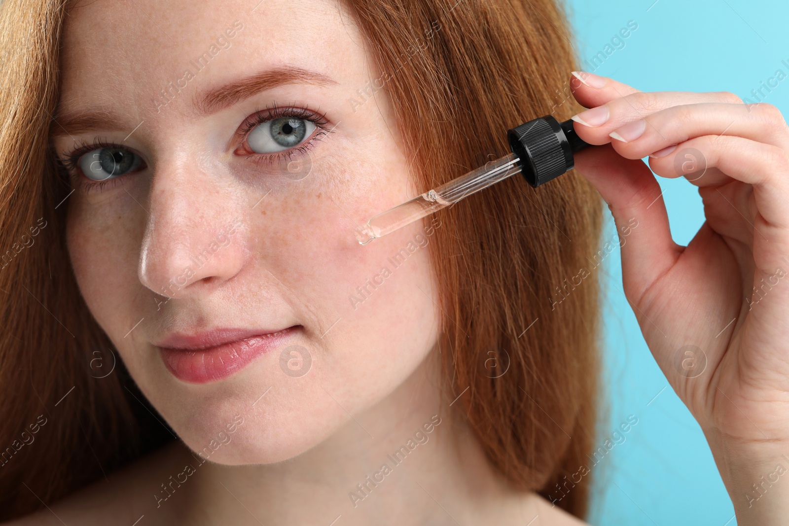 Photo of Beautiful woman with freckles applying cosmetic serum onto her face against light blue background, closeup