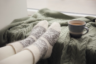 Photo of Woman wearing warm socks on knitted plaid near window, closeup. Cozy season