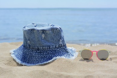 Photo of Jeans hat and sunglasses on sand near sea. Beach accessories