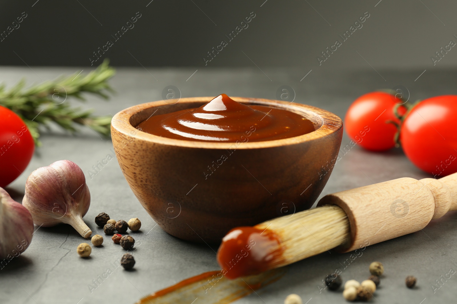 Photo of Tasty barbeque sauce in bowl, brush, garlic and peppercorns on grey table, closeup
