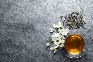 Glass cup of aromatic jasmine tea, dry leaves and fresh flowers on grey table, flat lay. Space for text