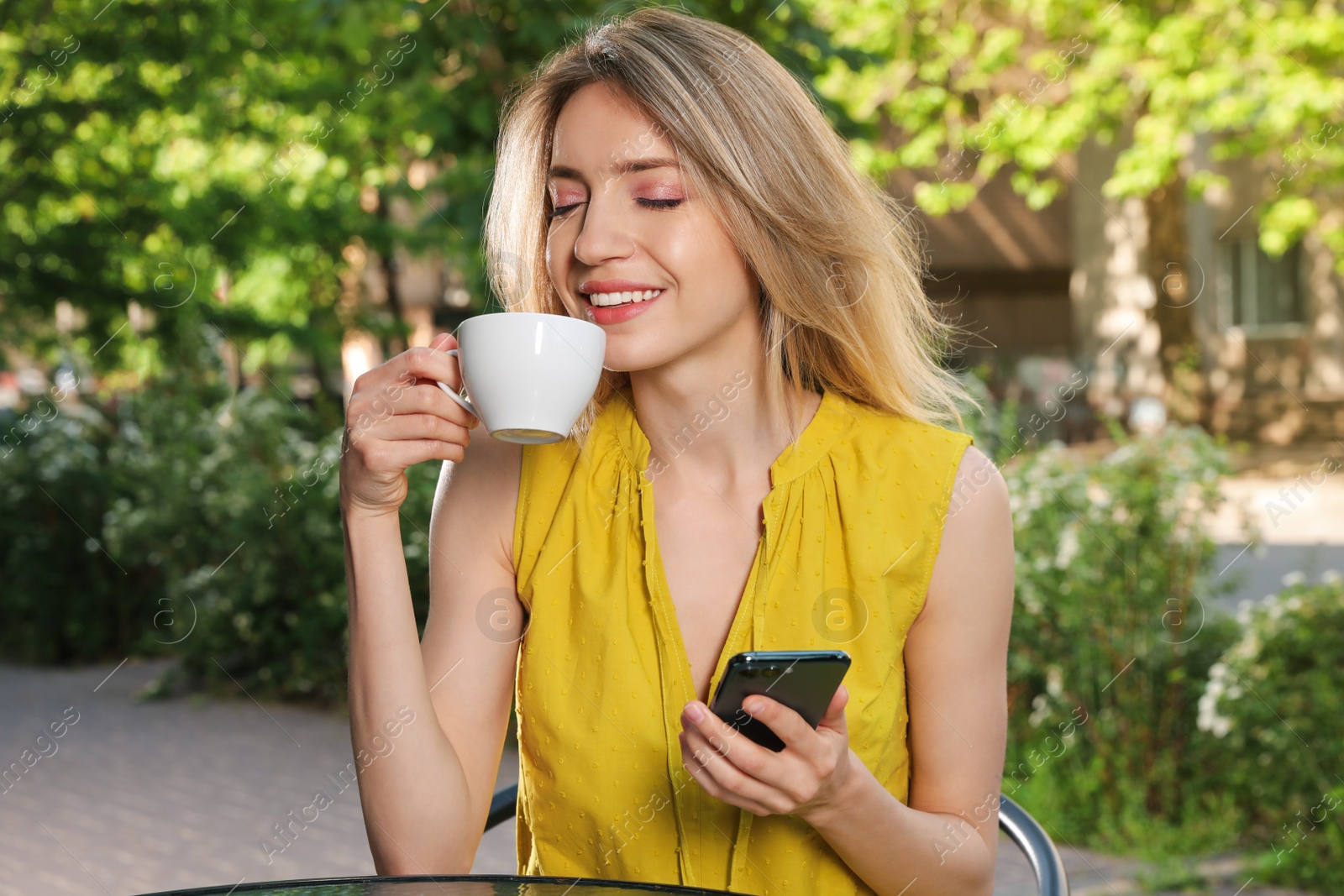 Photo of Happy young woman with cup of coffee and smartphone enjoying early morning in outdoor cafe